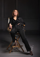 Beautiful adult woman sits on a stylish wooden director's chair on the black background. fashion model in black jacket.