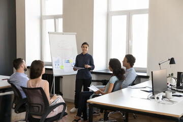 Diverse team meeting on office, listening to Indian project leader marketing report. Employee presenting financial charts to colleagues. Business coach, mentor woman teaching group of interns