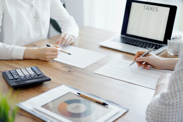 Woman accountant using a calculator and laptop computer while counting taxes with colleague at wooden desk in office. Teamwork in business audit and finance