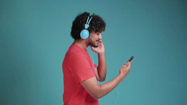handsome young indian guy in casual listen music on smartphone, using new headset, looking at cell phone and smiling, in coral t-shirt on blue studio background