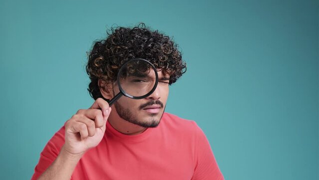 Funny swarthy man looking through magnifying glass, searching or investigating something, standing in coral t-shirt on blue studio background