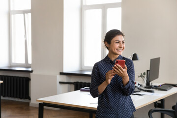 Happy dreamy young Indian business professional woman holding mobile phone, standing at work table in office, looking away, thinking over good news, chatting, smiling, laughing