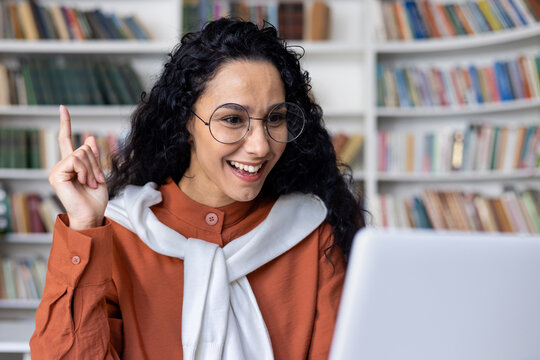 Idea concept. Excited young Indian woman wearing glasses sitting at a desk using a laptop and raising her index finger up in a classroom. Amazed, shocked lady, seeing, aha moment.