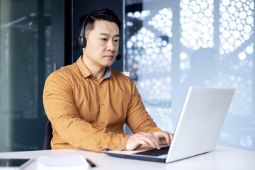 Concentrated Korean office worker in headset sitting at desk in front of laptop in modern office, Asian man working in call center advising customers, tapping on keyboard.