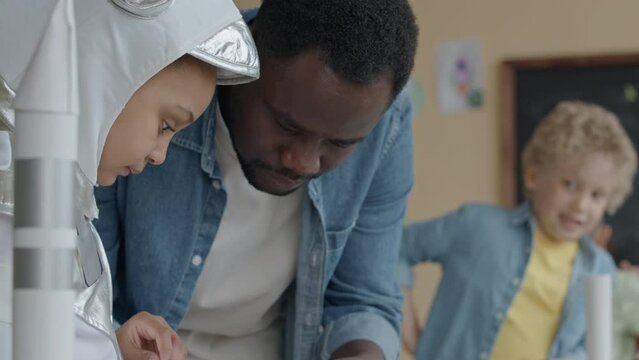 African American Teacher Using Glue While Helping Little Girl Dressed In Astronaut Costume To Make Space Rocket Out Of Paper During School Lesson