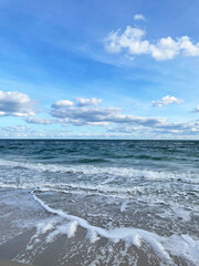 Seascape. Dark turquoise sea with small waves foaming near the shore, beige wet sand and a bright blue sky with white clouds. The horizon line is visible in the distance. Sunny autumn day.
