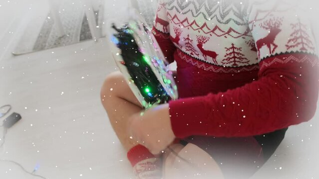 Close-up of man wrapping spool of glowing garland while removing decorations from a party until the next New Year. Sitting on the floor in red sweater.