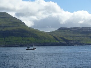 Beautiful scene of a boat in the sea near green mountains under blue sky in Faroe Islands