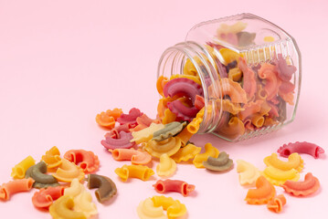 Glass jar with scattered colored variety formed pasta  on pink background