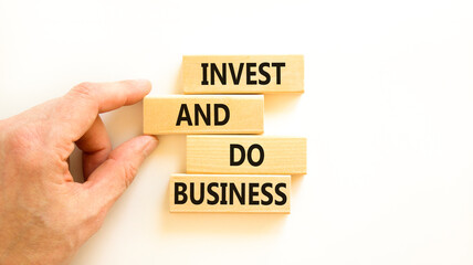Invest and do business symbol. Concept word Invest and do business on beautiful wooden block. Beautiful white table white background. Businessman hand. Invest and do business concept. Copy space.