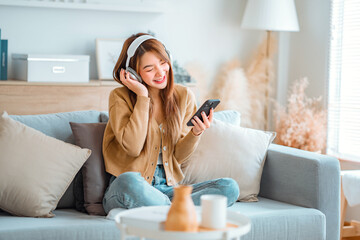 Happy asian woman listening to music from mobile phone while sitting on the the sofa at homes,...
