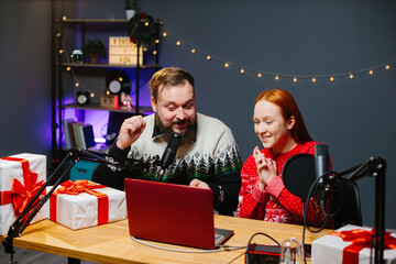 Christmas stream or video blog. Cheerful presenters in holiday clothes raffle off gifts for their subscribers. Male and female bloggers in New Year's outfits in a home studio.