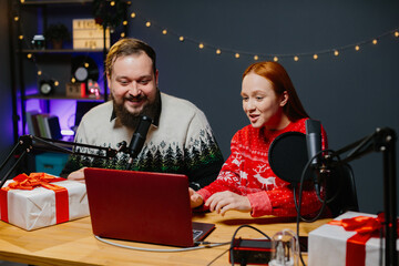 Christmas stream or video blog. Cheerful presenters in holiday clothes raffle off gifts for their subscribers. Male and female bloggers in New Year's outfits in a home studio.