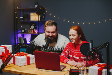 Christmas stream or video blog. Cheerful presenters in holiday clothes raffle off gifts for their subscribers. Male and female bloggers in New Year's outfits in a home studio.