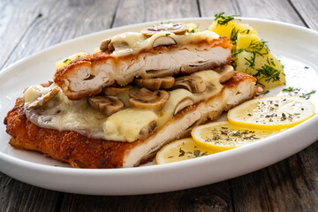 Crispy breaded seared chicken cutlet with fried white mushrooms, cheese and boiled potatoes on wooden table
