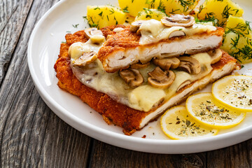 Crispy breaded seared chicken cutlet with fried white mushrooms, cheese and boiled potatoes on wooden table
