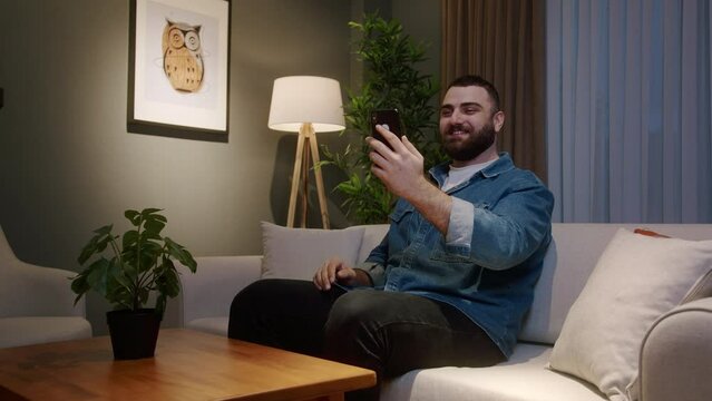 Young adult bearded man at home sitting on couch while having video call conversation through app with using smartphone, share news, talk to family living abroad, enjoy webchat virtual meeting
