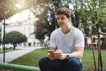 Obraz premium Young adult student using smart phone in park while going to school campus. Connected with friends everywhere, checking online bank, posting stories.