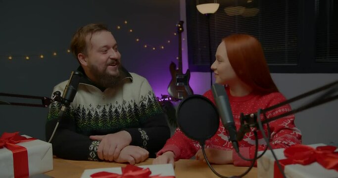 Christmas stream or video blog. Cheerful presenters in holiday clothes raffle off gifts for their subscribers. Male and female bloggers in New Year's outfits in a home studio.