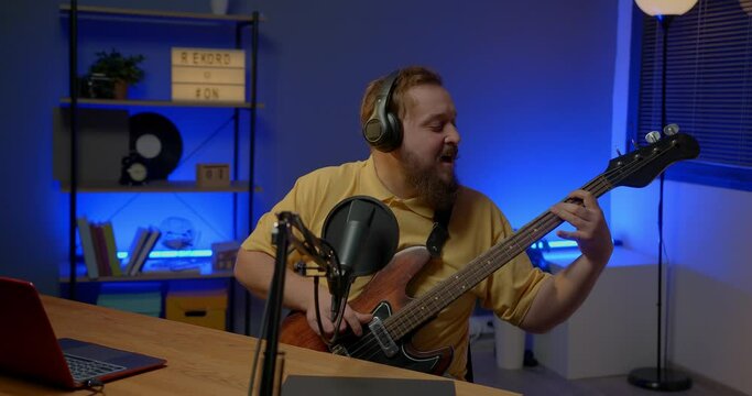 A man in headphones with a microphone on the table plays the bass guitar during an online broadcast. Online media or recording studio concept.