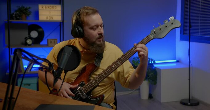 A man in headphones with a microphone on the table plays the bass guitar during an online broadcast. Online media or recording studio concept.