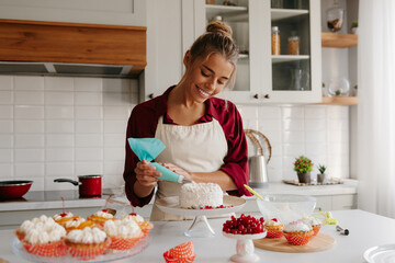 Joyful female baker applying whipped cream while decorating cake at the kitchen
