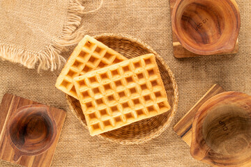 Two freshly baked aromatic waffles with straw plate and wooden cups on jute cloth, macro, top view.