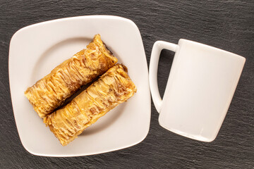 Two pieces of sweet baklava with white ceramic saucer and cup on slate stone, macro, top view.