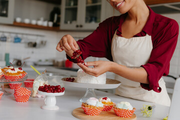 Close-up of female confectioner decorating cake with berries and smiling while standing at the kitchen