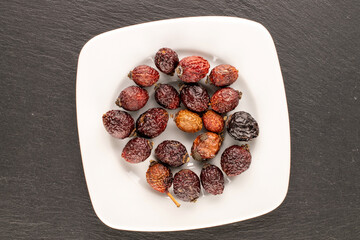Several dry rosehip berries with white ceramic plate on slate stone, macro, top view.