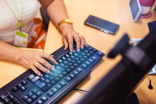 Senior Woman Working On A Computer, Her Hands In The Foreground; The PC Is Black In The Office. There Is A Blue Glow, And A Cell Phone Is Placed Next To The Keyboard.