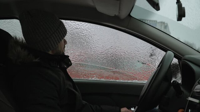 Man in a frozen car is surprised by ice outside window on cold winter day. Guy in warm clothes knocks out ice with his hand. Unhappy attitude towards winter weather and sub zero temperatures.