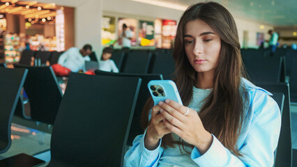 Portrait of young woman waiting airplane or train in waiting room with smartphone in hands. Beautiful woman typing on mobile phone at international airport waiting boarding at airport.