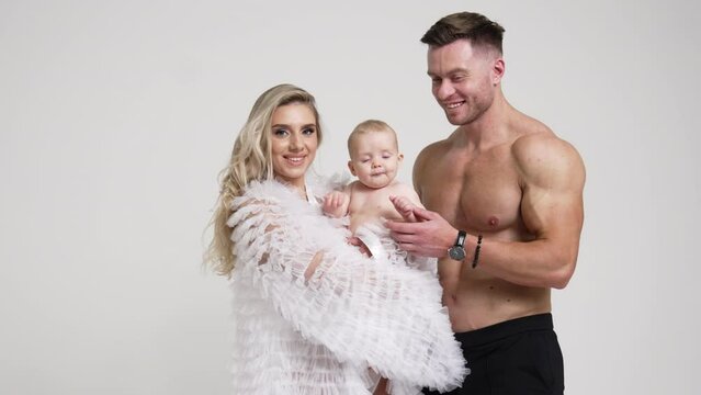 Lovely Caucasian couple with adorable infant baby. Muscular man with naked torso and his wife in white blouse holding a child. White backdrop.