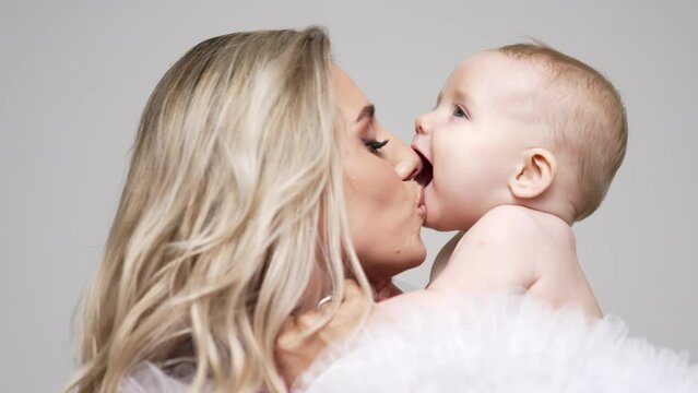 Loving mother with her beautiful infant baby. Blonde woman kissing her child and kid adorably opens his mouth. White backdrop.