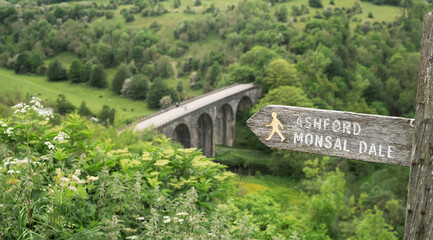 Footpath sign to Ashford Monsal Dale in front of the panoramic landscape looking down to the Monsal trail viaduct in Derbyshire Peak District. - 676944330