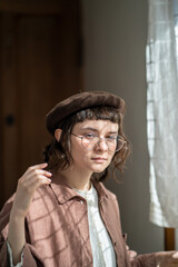 Melancholic teenager in vintage clothes and beret standing at window alone. Teen girl with sunlight shadow on face looks like rejected discriminated friend having difficulties in social communication