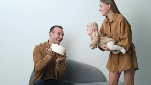 Joyful Caucasian parents amusing their little baby. Mother holds her child taking him closer to a cake in dad's hands.