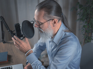 A male Presenter with a beard prepares to broadcast his audio podcast using a microphone and laptop...