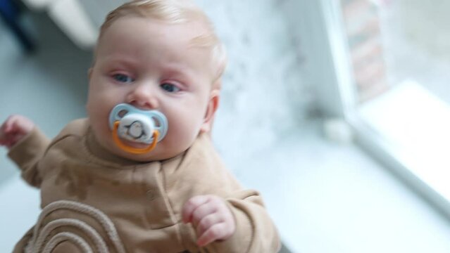 Cute blond baby boy with big blue eyes is waved in hands. Kid sucking a pacifier and looks at mom attentively.