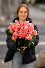 Fototapeta premium Young pretty dark-haired girl stands on a city street with a bouquet of pink roses