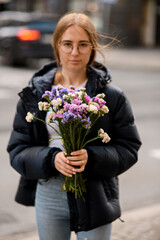Fototapeta premium Pretty girl with brown hairs stands on a city street with a bouquet
