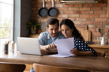 Young couple checking financial documents together, discussing loan agreement, sitting at table with laptop in kitchen, wife and husband calculating taxes, domestic bills, family planning budget - obrazy, fototapety, plakaty