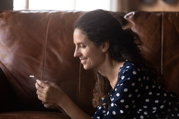 Side view smiling woman relaxing on couch with smartphone, looking at screen, writing message in...