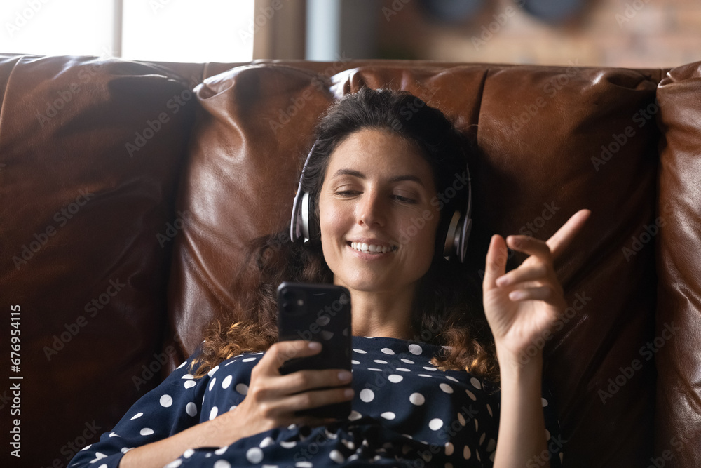 Wall mural Close up smiling woman in headphones listening to favorite music or podcast, using smartphone, relaxing on cozy couch at home alone, happy young female enjoying leisure time with gadget, having fun