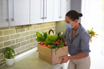 Young woman wearing medical mask, recyclable cardboard box with delivered fresh organic food on the...