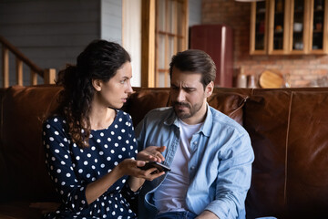 Unhappy jealous woman arguing with man, holding smartphone, sitting on couch at home, young family conflict, cheating in social networks and distrust concept, frustrated couple received bad news