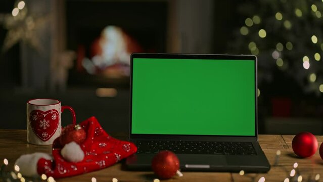 Laptop computer on desk at Christmas. Decorated room with Christmas tree and lights. Cosy winter home with fireplace. Blank green screen for web shop offer and holiday advertisement.