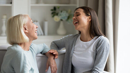 Obraz na płótnie Canvas Happy mature grandmother and granddaughter chatting, laughing at joke, smiling older mother and adult daughter having fun, talking and sitting on cozy sofa at home, sharing news, two generations