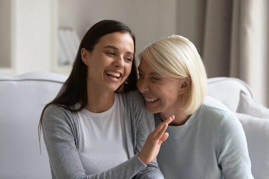 Happy young woman and mature mother chatting and laughing at joke, having fun at home together, sitting on cozy sofa, smiling positive grown daughter and senior mum talking, two generations